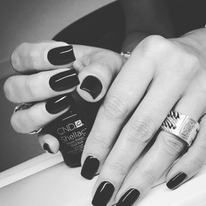 Zwart-witfoto van handen met zwarte nagels naast een flesje CND Shellac